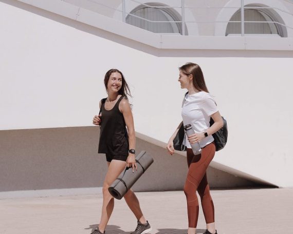 Two women in active wear walking outdoors while smiling and carrying yoga mats.