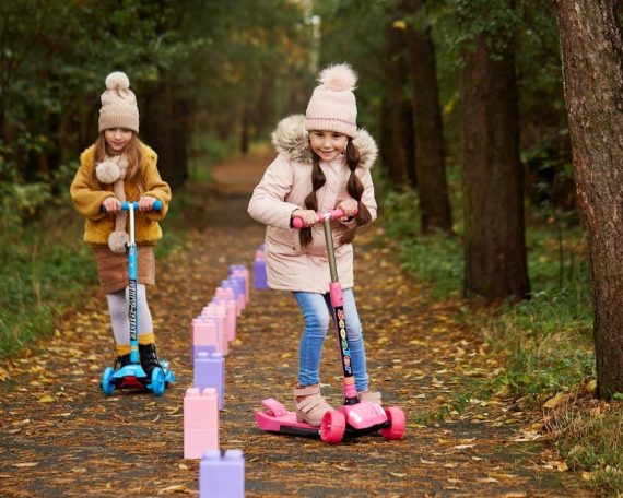 Two girls having fun on scooters in a leafy autumn park trail.